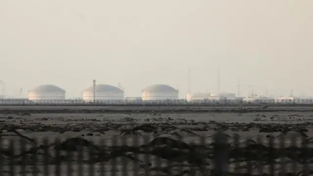 Several liquid gas tanks and electricity pylons are seen behind a fence in the distance on a hazy day