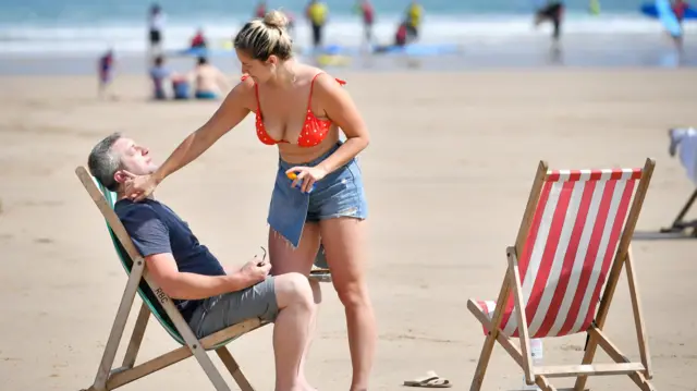 A woman in a bikini applies sunscreen to a man sat on a deckchair on a Cornish beach
