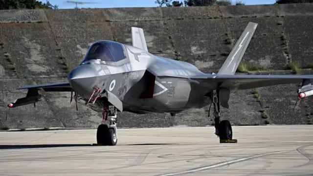 An F-35 jet, which is grey and has large wings, with wheels on the ground on tarmac.