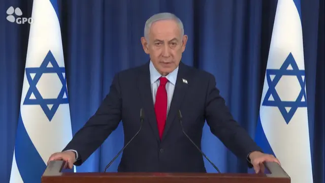 Bejamin Netanyahu leaning on wooden lectern as he delivers speech. He's standing in-between two Israeli flags