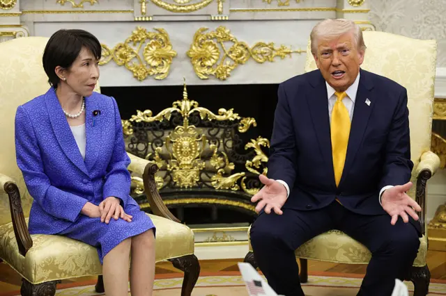 Trump and Japanese PM Takaichi sat on chairs in the Oval Office, gold chairs and backdrop, Trump has his palms open and facing the camera.