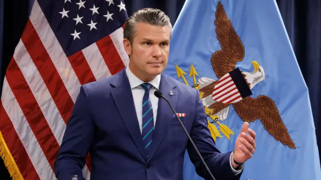 Pete Hegseth speaks into a microphone, standing in front of a US flag