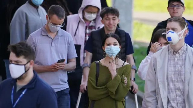 Several individuals are pictured outside with face masks on while queuing for a meningitis vaccine. The woman in the centre of the image is wearing a green jumper.