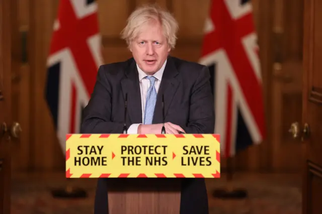Boris Johnson in front of a sign saying 'stay home, protect the NHS, save lives'