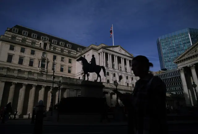 Shot of the exterior of the Bank of England building on a sunny day. A statue of a soldier on a horse stands on top of a stone plinth in front of the building, in the foreground is a woman on her phone, in the backlight