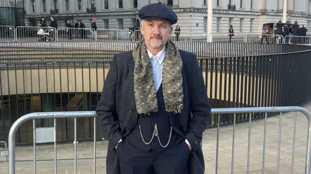 A man wearing a black flat cap, two piece suit with a gold chain on the waistcoat and a brown and green scarf. He has his hands in his pockets and a wide stance. He is standing in front of metal barriers