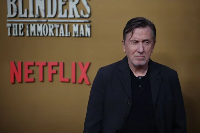 A man with brown and grey hair in a black shirt and suit jacket in front of a gold backdrop. He is winking at cameras