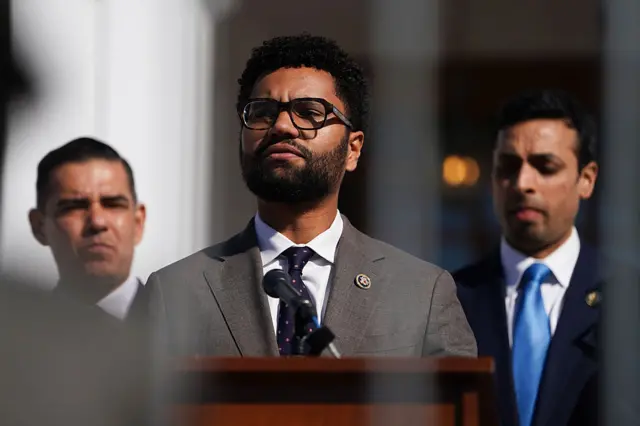 Maxwell Frost speaks after Clinton deposition, in a suit and drak rimmed glasses, at a podium, flanked by two other lawmakers