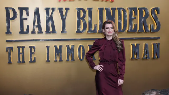Karen Carney is in a maroon silk dress and has long brown hair. She is standing in front of a gold backdrop with black writing on it that reads "peaky binders the immortal man".