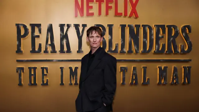A man with brown hair is in an all black suit. He is standing in front of a gold film premiere background with black writing that reads "peakyblinders the immortal man"