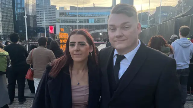A young man and woman. The man has light brown hair swept to the side and is wearing a black suit jacket, tie and waistcoat and a white shirt. The woman has long red hair, a blush pink dress and black jacket
