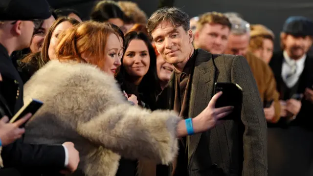 A woman with ginger hair and a brown fur is holding out a black phone and Cillian Murphy is in a brown shirt and black suit jacket, posing with her