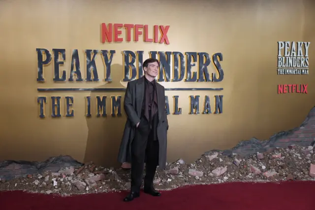 Cillian Murphy has a dark brown silk shirt on with a long black coat. He is smiling and standing in front of a large gold backdrop that reads "peaky blinders, the immortal man".