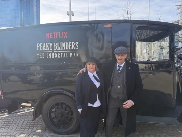 Claire White and Albert Laszlo wearing Peaky Blinders-style clothing standing in front of a promotional van