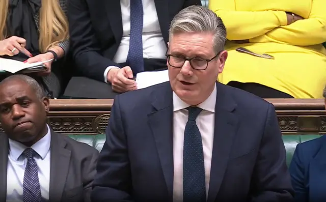 Keir Starmer addressing MPs in the Commons. He's in a dark suit, sitting on green leather benches to his right is deputy prime minister David Lammy