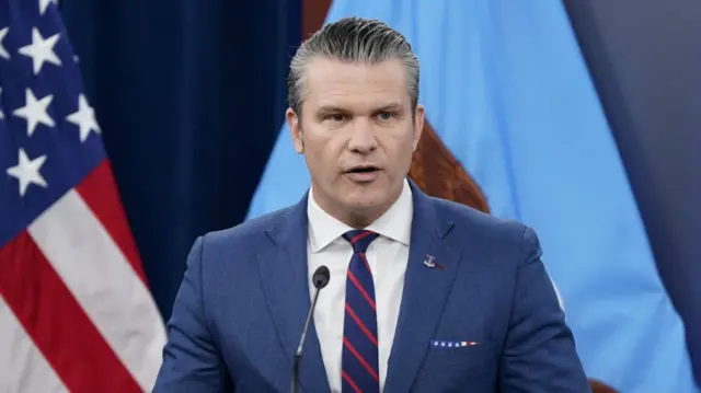 Pete Hegseth in blue-greyish suit and striped tie speaks at the Pentagon. A US flag to his right