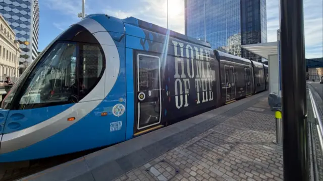 A tram vehicle at a station that has a blue drivers cab. The rest of the tram is black and brown, with large white writing that reads "Home of the". On the cab it says "Steven Knight" which is the tram's name