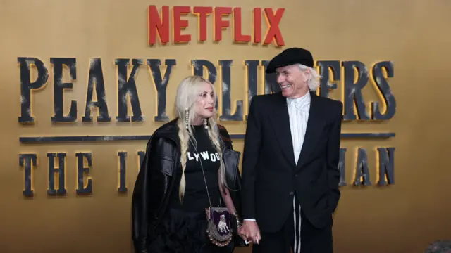 A man and woman both with white blonde hair are on a red carpet in front of a gold backdrop with writing that says "peaky blinders the immortal man". The man is in a black jacket and trousers with a white shirt and black flat cap while the woman is in a black tshirt with white writing on it, a black jacket and a small bag with feathers on it
