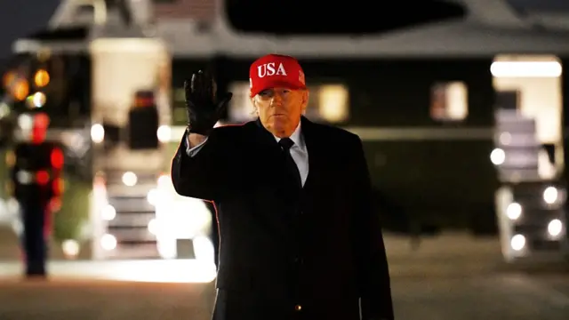 An elderly man in a dark coat, gloves and red baseball cap raises his hand in front of an aeroplane at night time