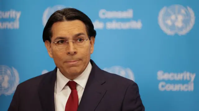 Danny Danon in black suit, white shirt and red tie stands in front of a wall covered in a blue paper with the UN Security Council logo