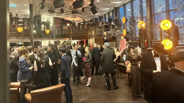 A room full of people with brown wooden floors and an upstairs balcony area. People are dressed like Peaky Blinders with flat caps and suits on, as well as sparkly dresses