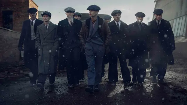 Production still from The Immortal Man showing eight male cast members wearing flat caps, suits and overcoats, walking down a Birmingham street