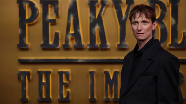 A man with brown hair in a black suit standing in front of a gold backdrop with large words saying "peaky blinders" on it