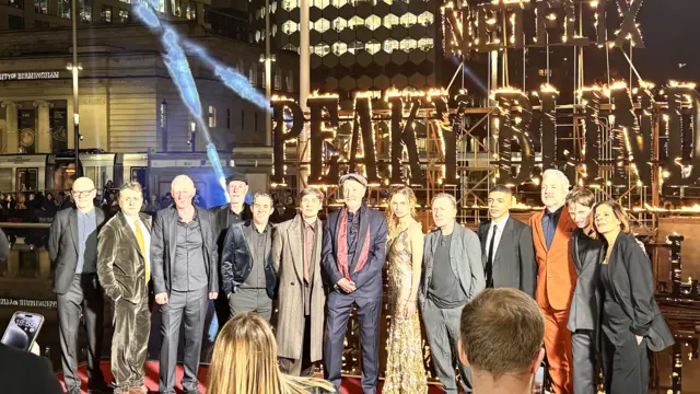 13 people standing in front of a huge sign that reads "Peaky Blinders" which is engulfed in flames. Eleven men are in suits with one woman in a gold dress and another in a black suit.