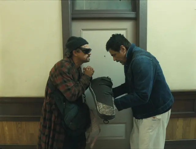 Leonardo DiCaprio and Benicio del Toro standing over a violin case in a hallway