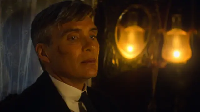 Tommy Shelby has grey and brown hair which sweeps to the side. He is wearing a suit jacket with a white shirt in a dimly lit room. He has a crescent shaped cut on his cheek with dried blood on it. On a wall next to him is a gold mirror with a hanging lamp which illuminates Shelby's face in a soft yellow glow.