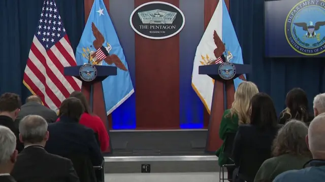 Reporters sitting in front of a stage with two lecterns bearing the US sigil in the Pentagon