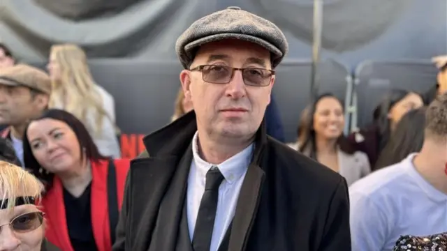 A man with a grey flat cap, tinted glasses, a white shirt, black tie and waistcoat and a black jacket