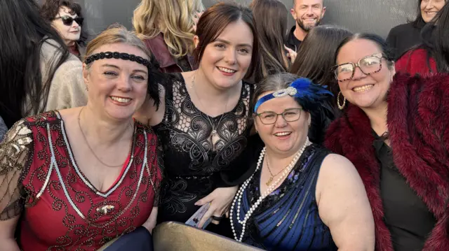 Four women are posing for a photo together at the film premiere. On the far left the woman is blonde with a red and black dress with sequin detailing and a black sequin and feather headband. Next to her a woman has brown hair tied back and a black dress with swirl pattern detailing. To her right is a woman with brown hair tied back, a blue and black sequin dress and a blue feather headband. On the far right is a woman with black hair, glasses and a black dress with a red fur jacket