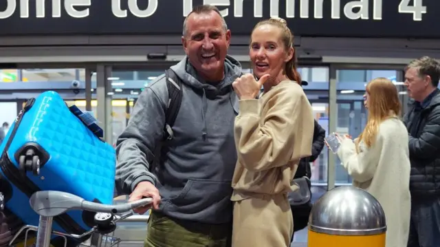 Outside Terminal 4, a couple smile at the camera while holding their luggage, with the woman holding her thumbs up
