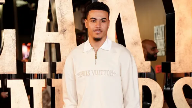 A man with black hair and black facial hair is wearing a cream jumper that says "louis vuitton" on it and is standing in front of large gold letters that form part of the words "peaky blinders"