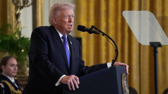 Donald Trump mid-speech as he leans with both hands onto a lectern inside the White House's East Room
