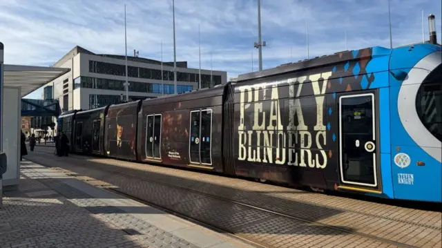 The other half of the metro tram, which is brown and has Tommy Shelby on the front, with much of the top of his face covered by a flat cap. In white writing it reads "Peaky Blinders" and says "Steven Knight" on the blue half of the back cab