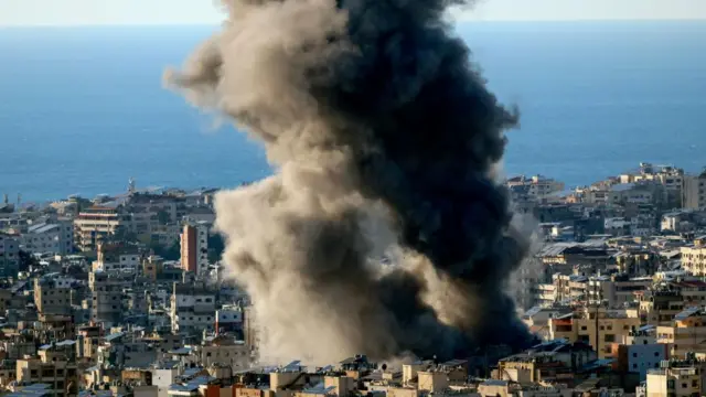 Large plumes of smoke rise from buildings, with the sea just behind