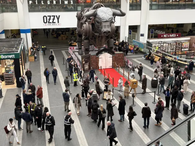 Ozzy the bull on the concourse of New Street Station. There is a red carpet in front of him leading to two white gates with commuters surrounding him, many taking photographs