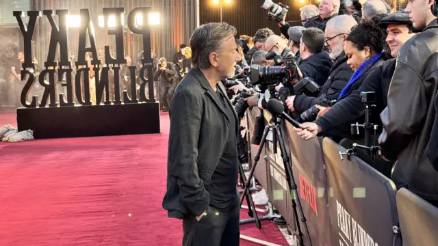 A man in a black suit and t-shirt speaking to journalists who are holding onto microphones. There is a large sign that reads "peaky blinders" and a red carpet