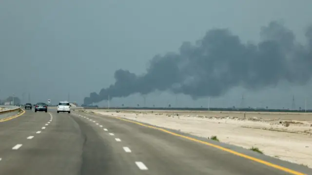 Smoke billows from the Saudi Ras Tanura oil refinery, cars drive on a road nearby.