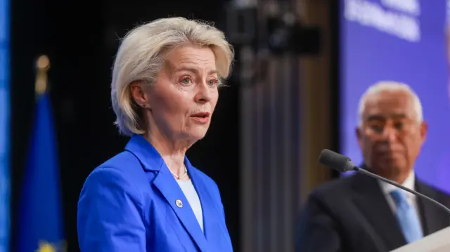European Commission President Ursula von der Leyen speaks from behind a podium, wearing a royal blue jacket and a pearl necklace.