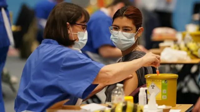 A student receiving an injection in the sports hall at the University of Kent campus in Canterbury. A medical professional in scrubs and the student are both wearing masks