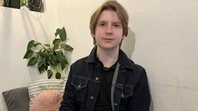 A teenage boy with blond, shaggy hair and a black jacket sitting against a white wall.