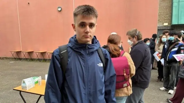 Young man in dark blue rain jacket poses for camera in front of a small wooden table, people with face masks queueing outside a red building behind him