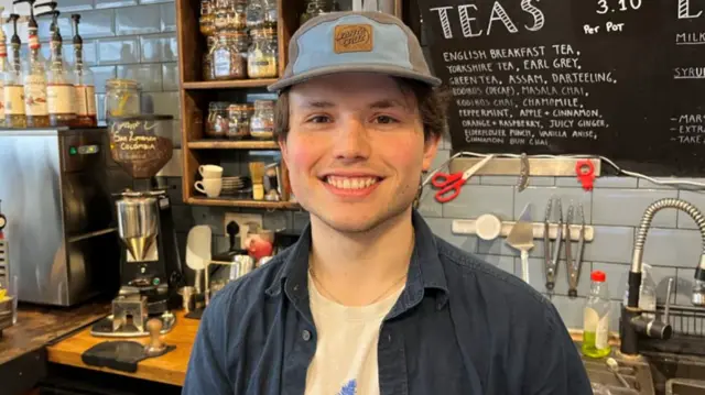 A young man wearing a cap and navy open shirt smiles while standing in a cafe with a coffee machine in the background.