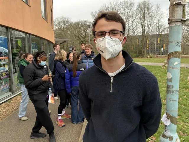 Young man in a black hoodie poses for pic, He's wearing glasses and a face mask, he's standing in a queue outside on the side of a building