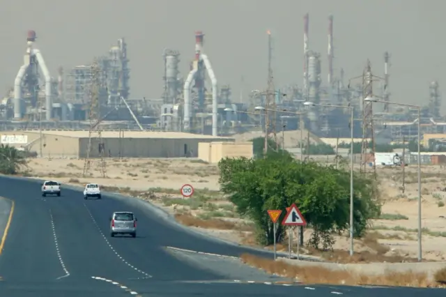A distant shot of the refinery, showing lots of chimneys, with some cars travelling on a road closer to the camera.