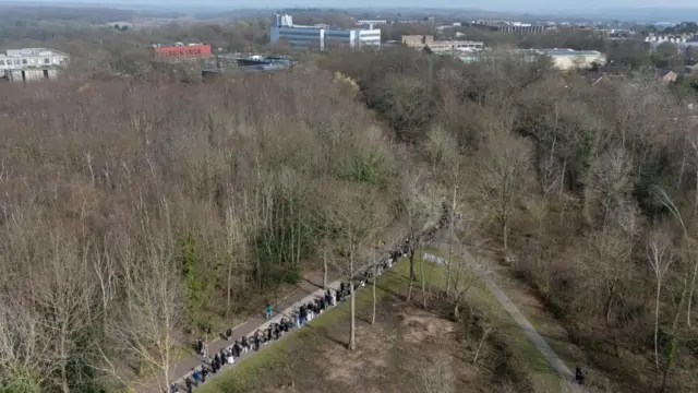 An aerial view that shows a long queue of people standing on a footpath next to a road that cuts through a forested area.