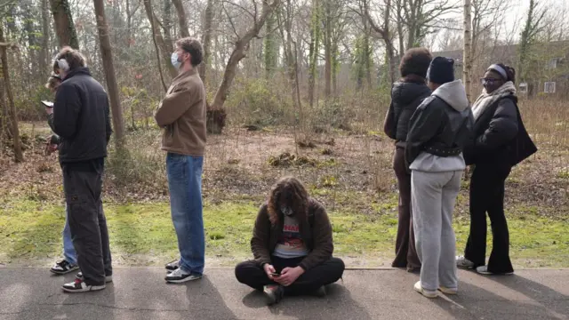 A student sits on the ground as others stand in line either side of them along a tree-lined street.